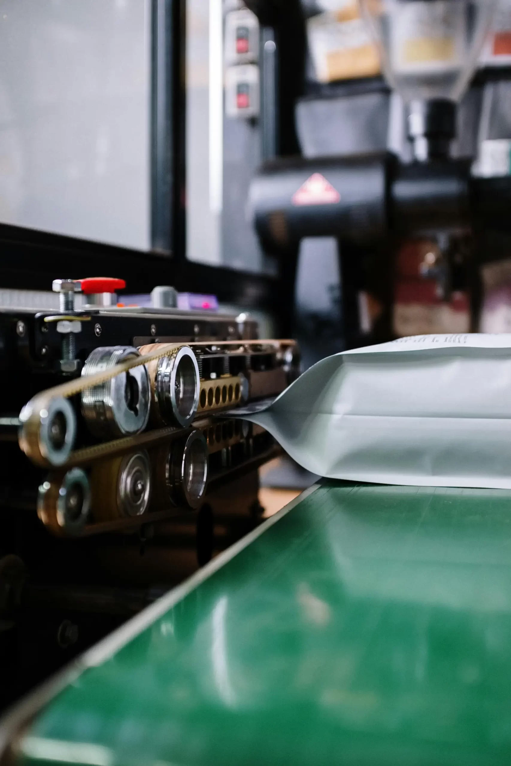 An industrial continuous band sealer machine sealing a silver foil coffee bag on a green conveyor belt in a production facility